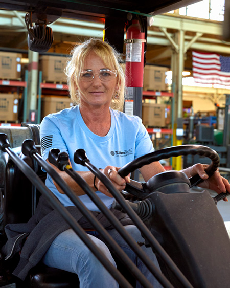 A woman wearing safety glasses and notably a TimberTech t-shirt pauses to pose for a photo while driving a forklift